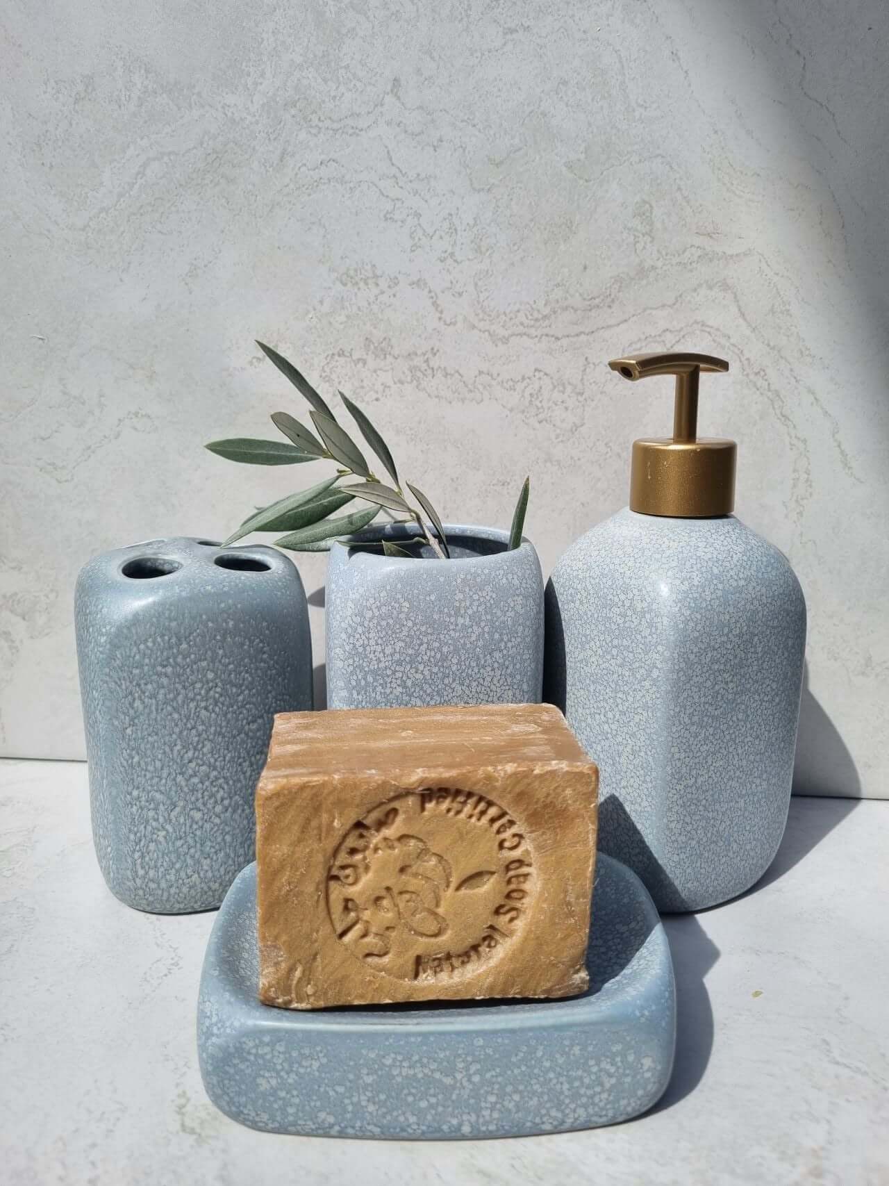 Elegant blue bath accessory set with soap dispenser, toothbrush holder, and soap dish, featuring natural soap and greenery.