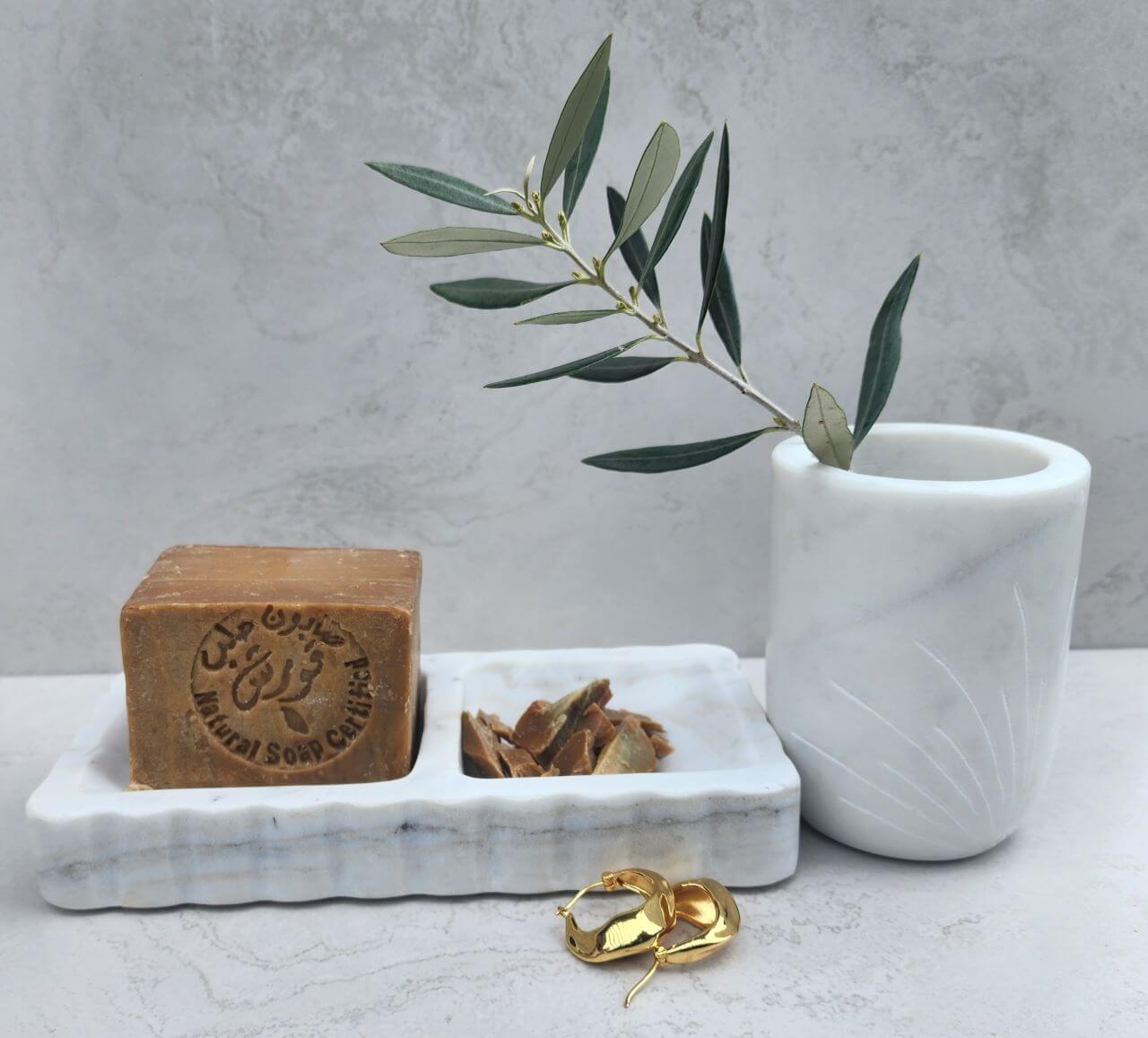 Marble soap dish holding bar soap alongside a matching marble tumbler with greenery, set on a textured bathroom surface.